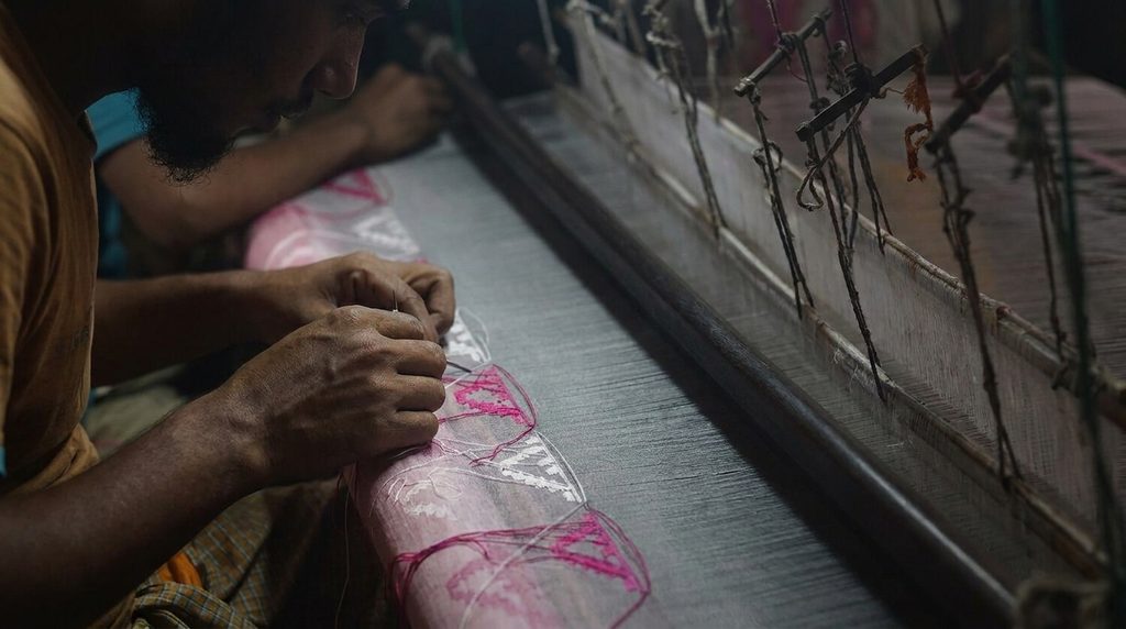Close-up of a Jamdani weaver's hands threading pink and white floral motifs onto fabric using a needle on a traditional wooden handloom
